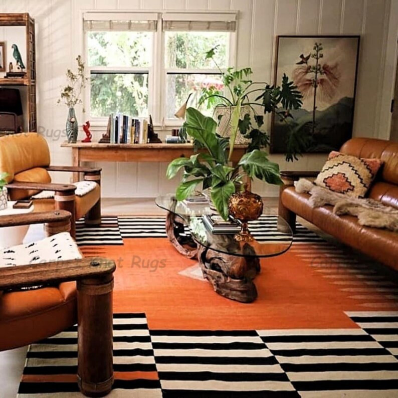 Op de afbeelding: Een woonkamer met een feloranje tapijt met zwart-witte geometrische patronen. De kamer omvat leren stoelen en een bank, een glazen salontafel en een houten boekenplank. Een groot schilderij en planten dragen bij aan de inrichting.