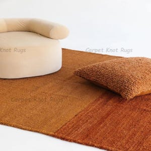 May include: A warm-toned, textured rug with a two-tone design. A beige, rounded chair and a brown, textured pillow are placed on the rug. The rug has the text "Carpet Knot Rugs" printed on it.