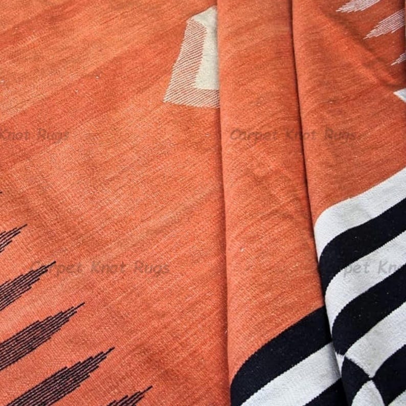 Op de afbeelding: Close-up van een opgevouwen oranje tapijt met zwart-witte gestreepte accenten. Het tapijt heeft een getextureerd oppervlak en een geometrisch ontwerp. Het tapijt is gemaakt van een geweven materiaal.