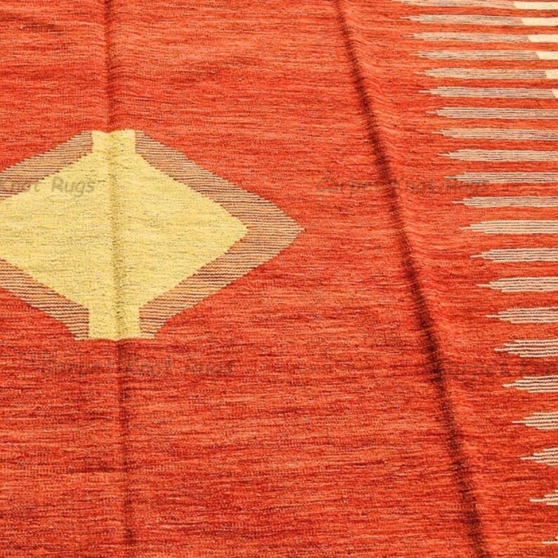 Op de afbeelding: Close-up van een getextureerd, oranje tapijt met een geel ruitvormig ontwerp in het midden. Het tapijt heeft een streepjespatroon aan de rechterkant en de tekst "e Rugs" in de linker benedenhoek.