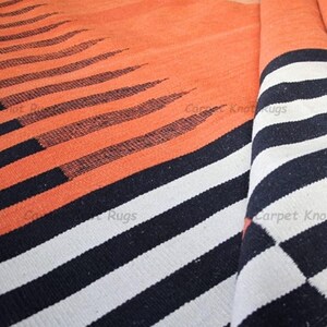 Op de afbeelding: Oranje en zwart gestreept tapijt met een opgerold gedeelte met zwart-witte strepen. Het tapijt heeft een textuur, met het oranje gedeelte met zwarte lijnen die taps toelopen.