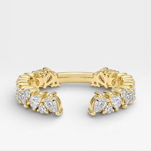 May include: A gold-colored ring with a unique open design. The ring is adorned with multiple clear, sparkling gemstones. The gemstones are clustered together, creating a dazzling display. The ring is set against a plain white background.