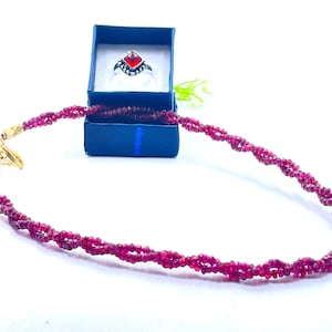 Può includere: Una collana di rubini con chiusura dorata e un anello abbinato in una scatola blu. L'anello ha una fascia argentata e una pietra preziosa rossa. La collana e l'anello sono esposti su una superficie bianca.