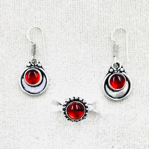 Può includere: Un set di gioielli con un anello e un paio di orecchini. Ogni pezzo ha una montatura in metallo color argento con una gemma rotonda rossa vibrante. Gli orecchini sono a goccia e l'anello ha un design floreale attorno alla gemma.