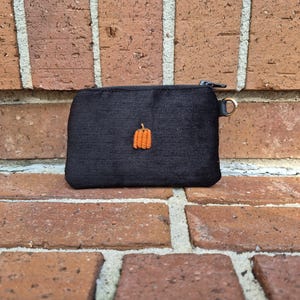 May include: A small, rectangular black zippered pouch made of a textured fabric. The front of the pouch features a single, small, embroidered orange pumpkin icon centered on the surface. A small metal ring is attached to the side zipper pull.