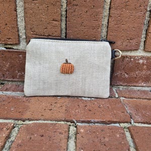 May include: A rectangular beige linen coin purse featuring a small, embroidered orange pumpkin centered on the front. The pouch includes a black zipper closure along the top edge and a small gold-toned metal ring attached to the side for hanging.