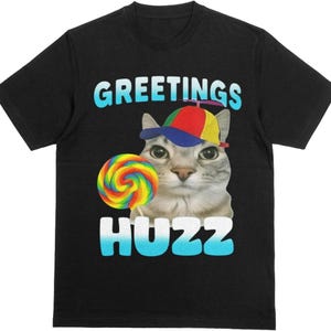 May include: A black t-shirt featuring a graphic of a cat wearing a multicolored propeller hat. The cat is holding a rainbow-colored lollipop. The text GREETINGS is printed in light blue above the cat, and the word HUZZ is printed in white below.