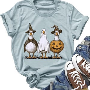 May include: A light blue t-shirt featuring a graphic of three white geese dressed for Halloween. One goose wears a witch hat and cape, the middle goose is covered by a white ghost sheet, and the third goose wears a witch hat and a carved pumpkin.