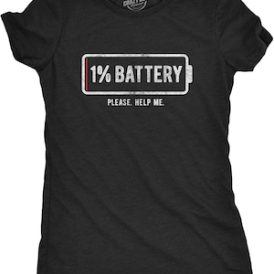 Pode incluir: Uma t-shirt preta de manga curta com um gráfico branco de um ícone de bateria fraca com uma barra indicadora vermelha. O gráfico inclui o texto 1% BATTERY e a frase PLEASE. HELP ME. por baixo. A t-shirt tem gola redonda e mangas curtas.