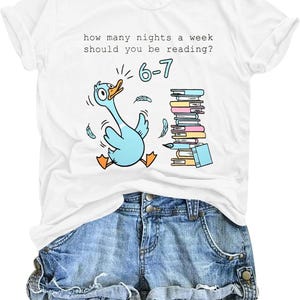 May include: A white t-shirt featuring a graphic of a light blue duck with orange feet and beak, standing next to a stack of colorful books. The text reads, "how many nights a week should you be reading? 6-7". The shirt is paired with blue denim shorts.