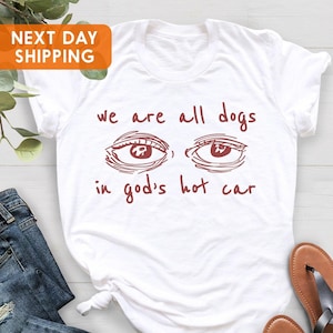 May include: A white short-sleeved t-shirt featuring a graphic of two eyes in the center. Below and above the eyes, the text reads: we are all dogs in god's hot car. The graphic and text are printed in a dark reddish-brown color on the fabric.