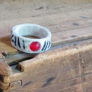 以下が含まれることがあります： 黒い縞模様と鮮やかな赤い宝石が付いた白い指輪。素朴な木製の表面に置かれた指輪は、そのユニークなデザインと職人技を際立たせています。指輪のデザインは目を引きます。