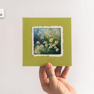 Op de afbeelding: Een handgeschilderde aquarel van madeliefjes op een groene kaart. Het kunstwerk toont witte madeliefjes met gele centra tegen een groene en blauwe achtergrond. De kaart heeft een gescheurde rand. Tekst op de kaart luidt "Hand Painted Original Watercolor."