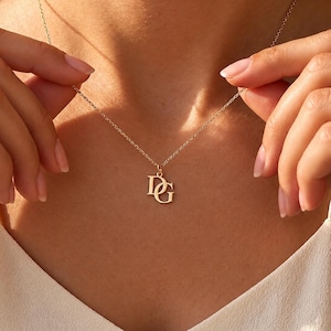 May include: A gold-toned necklace featuring a delicate chain and a pendant with the letters D and G in a serif font. The pendant hangs centrally from the chain, which rests against the collarbone area, showcasing a polished metallic finish.