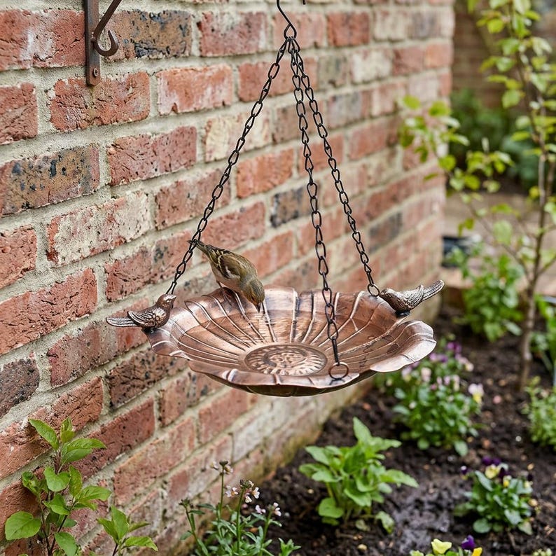 May include: A copper-colored metal bird bath with a scalloped edge and a central floral pattern. The basin is suspended by three black metal chains attached to small decorative bird figures on the rim, with a small bird perched inside.