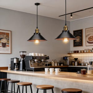 May include: Two black pendant lights with copper-coloured interiors hang above a coffee shop counter. The lights have a vintage industrial design with exposed Edison-style bulbs. The shop features a coffee machine, grinder, and stools.