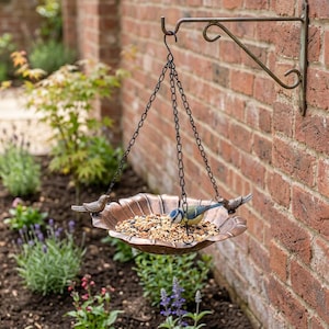 May include: A copper-colored metal bird feeder hanging from a wall-mounted bracket by three black chains. The circular, scalloped-edge tray is filled with birdseed and features two small decorative bird figures attached to the rim.