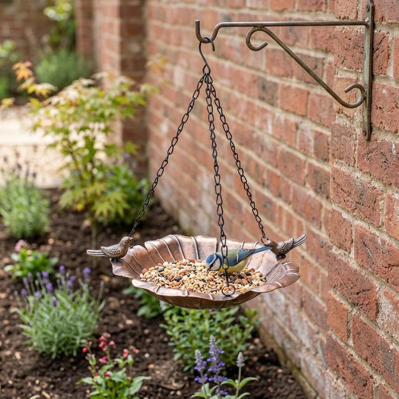 May include: A copper-colored metal bird feeder hanging from a wall-mounted bracket by three black chains. The circular, scalloped-edge tray is filled with birdseed and features two small decorative bird figures attached to the rim.