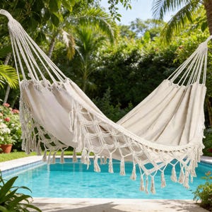 Puede incluir: Una hamaca color crema con detalles de macramé y flecos cuelga sobre una piscina. La hamaca está suspendida de cuerdas y está rodeada de exuberante vegetación, creando una escena relajante al aire libre.