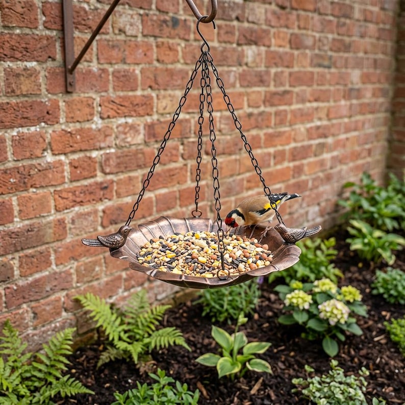 May include: A hanging bird feeder featuring a shallow, scalloped, copper-toned metal bowl suspended by three dark metal chains. Two small, decorative metal birds are attached to the rim of the bowl, which is filled with a variety of birdseed.