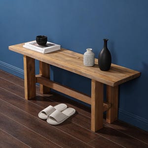 May include: A wooden bench with a natural finish, featuring a black ceramic bowl, books, two vases, and light grey slippers. The background is a dark blue wall and a dark brown wooden floor. The bench is a stylish piece of furniture.