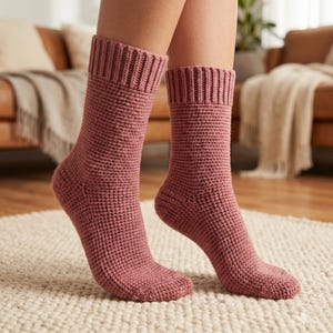 May include: Mauve knitted socks with ribbed cuffs. The socks have a textured, woven appearance and are shown on a light-colored rug. The socks are a warm, cozy style.