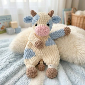 Puede incluir: Un peluche de vaca de ganchillo hecho a mano en crema, azul claro, rosa y marrón. La vaca tiene manchas azules, una nariz rosa y pezuñas y cuernos marrones. El juguete está sobre una superficie blanca y esponjosa y una manta de punto azul claro.