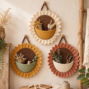 May include: Three circular crochet wall hangings shaped like sunflowers. Each piece features a dark brown center, a semi-circular pocket, and textured petals in cream, yellow, and terracotta. The pockets hold dried flowers, greenery, and scissors.