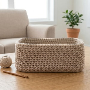 Könnte beinhalten: Ein rechteckiger, beigefarbener Häkelkorb steht auf einem Holztisch. Eine Garnkugel und eine Häkelnadel aus Holz liegen neben dem Korb. Ein beiges Sofa und eine Topfpflanze sind im Hintergrund.