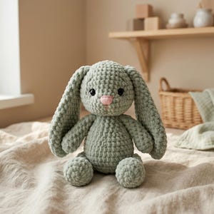 May include: A sage green crochet bunny toy with long ears, a pink nose, and black eyes. The soft toy is sitting on a beige fabric. The handmade bunny is a cuddly toy.
