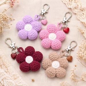 May include: Four crochet flower keychains in lavender, pink, burgundy, and beige. Each flower has a white center and is attached to a silver-toned metal lobster clasp with a small, solid-colored heart charm dangling from the ring.