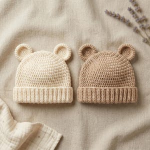 Può includere: Due cappellini da neonato all'uncinetto, color crema e beige, ciascuno con orecchie a forma di orso. I cappellini hanno un bordo a coste e un punto strutturato per la corona. I cappellini sono esposti su una superficie di lino neutra.