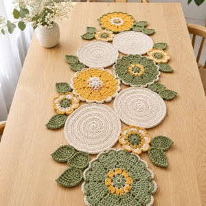 May include: A handmade crochet table runner featuring a floral design. The runner includes yellow, green, and cream-colored flowers and leaves. The runner is laid out on a light-colored wooden table.