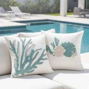 Peut inclure: Deux coussins blancs avec des motifs de récifs coralliens brodés en teintes de turquoise et beige. Les coussins sont placés sur un canapé extérieur blanc près d'une piscine, créant un thème côtier.