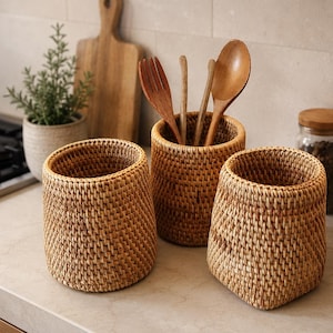 Könnte beinhalten: Drei gewebte Utensilienhalter aus Rattan in verschiedenen Größen stehen auf einer Arbeitsplatte. Einer enthält Holzlöffel und eine Gabel. Eine kleine Topfpflanze und ein Holzschneidebrett befinden sich im Hintergrund. Gläser mit Holzdeckeln sind ebenfalls sichtbar.