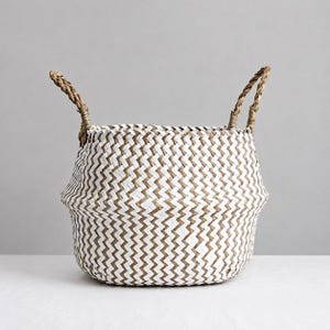 Può includere: Un cesto intrecciato con un motivo a zig-zag bianco e marrone. Il cesto ha due manici intrecciati e una forma svasata. Il cesto è realizzato con materiali naturali ed è adatto per riporre oggetti o decorare.