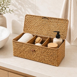 May include: A rectangular woven storage basket with a hinged lid, divided into four compartments. The basket contains a white towel, a brush, a small wooden container, and a lotion bottle. The basket is made of a light brown material.