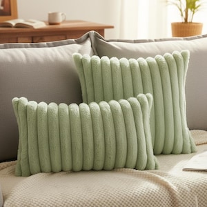Puede incluir: Dos cojines decorativos verde salvia con textura acanalada. Un cojín es rectangular y el otro cuadrado. Los cojines están sobre un sofá gris claro con una manta color crema.
