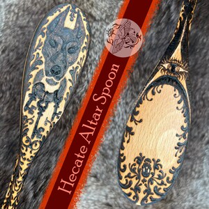 May include: Two wooden altar spoons with intricate carvings. One spoon features a wolf-like figure and keys, while the other displays an eye and floral patterns. The text "Hecate Altar Spoon" is visible on a red stripe.
