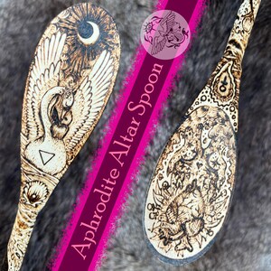 May include: Two wooden altar spoons with intricate pyrography designs. One spoon features a swan with a crescent moon and sunburst, the other a detailed mythological scene. The text "Aphrodite Altar Spoon" is visible.