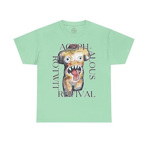May include: A light green t-shirt featuring a watercolor illustration of a cartoon dog with an open mouth and a long tongue. The text "ACEPH-ALOUS ROTWIT REVIVAL" is printed on the shirt in black.