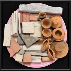 May include: A collection of miniature terracotta bowls, lamps, and stone tiles in shades of grey, beige, and red. The items are arranged on a pink and white striped plate. The background is black.
