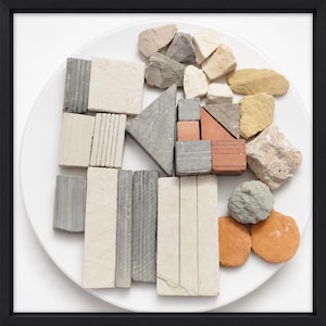Può includere: Una collezione di pietre e pezzi di mattoni di varie dimensioni e colori, disposti su un piatto bianco. Le pietre includono sfumature di grigio, beige, arancione e marrone. La disposizione suggerisce un tema di blocchi di costruzione.