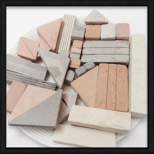 May include: An assortment of small, geometric stone blocks in various shapes and colors, including triangles, rectangles, and squares. The blocks are in shades of gray, beige, and peach, arranged on a white, ridged plate.