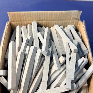 Peut inclure: Une boîte en carton remplie de nombreux bâtonnets de craie gris hexagonaux. Les morceaux de craie sont de longueurs variables et sont disposés au hasard. La boîte est ouverte, révélant le contenu.