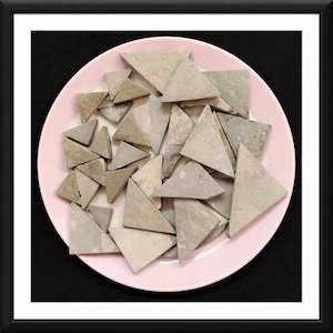 May include: A collection of gray, triangular stone tiles arranged on a light pink plate. The tiles vary in shades of gray and beige. The plate is centered against a black background, creating a contrast that highlights the tiles.