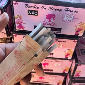 Könnte beinhalten: Eine Packung grauer Schieferstifte mit einer braunen Papierhülle, beschriftet mit "Barbie Slate Pencil". Der Hintergrund zeigt rosa Schachteln mit dem Text "Barbie In Every House" und Barbie-Illustrationen. Die Schachteln haben auch die Buchstaben "ABC".
