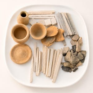 May include: A collection of natural clay items including a small pitcher, two bowls, a saucer, several rectangular clay sticks, small cubes, and irregular stone-like fragments arranged on a white plate. The items are in shades of tan, gray, and white.