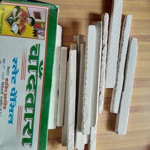 May include: A rectangular green and white box labelled Chandtara containing multiple thin, off-white slate pencils. The box features a graphic illustration of a person reading a book. The pencils are arranged in a loose, vertical cluster next to the box.