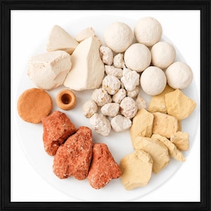 May include: A white plate displays various natural clay products. There are large, irregular chunks of beige and red clay, along with smaller, pebble-like pieces. Several smooth, round clay balls and a small, terracotta-colored pot are also present.